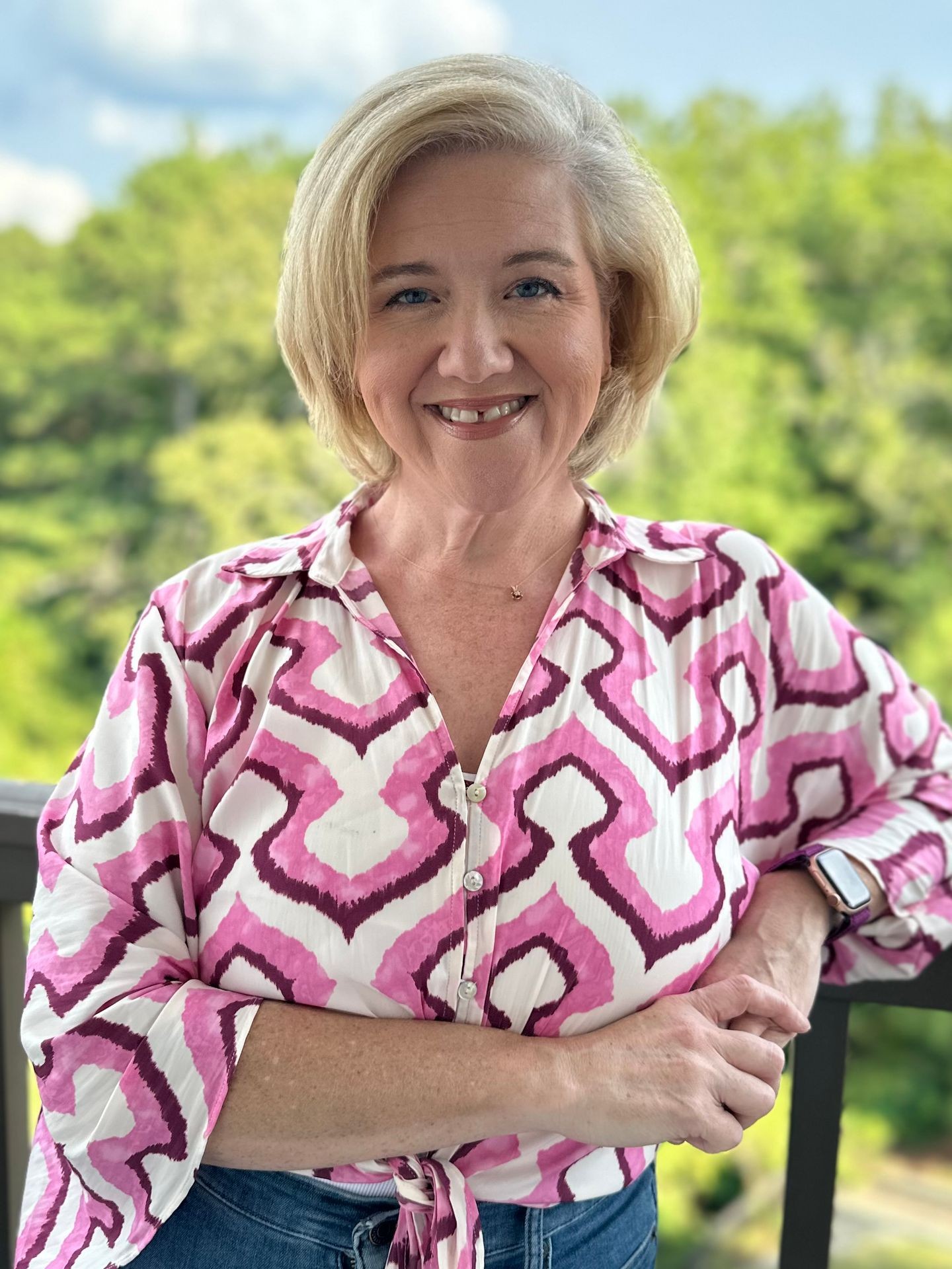 Angela, founder of Oxford Organized, smiling and standing on a balcony in a pink and white patterned blouse with a scenic background of trees and sky.